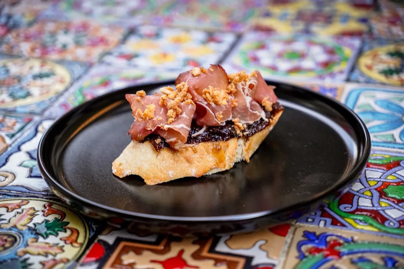 Pintxo na černém talíři na malovaných kachličkách.